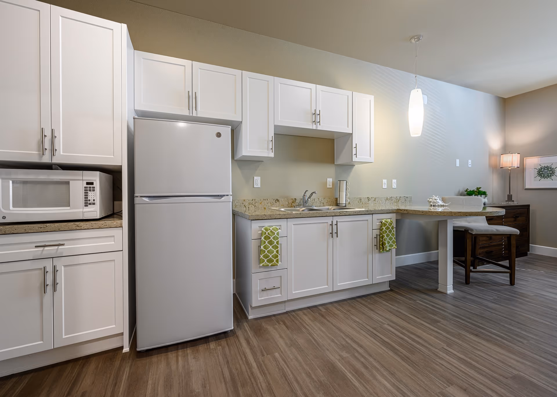 A modern kitchen area with white cabinets, a white refrigerator, a microwave, and a granite countertop with a sink. Two green patterned towels hang from the cabinet doors. The kitchen extends into a small dining area with a wooden chair, a pendant light, a table lamp, and a framed botanical print on the wall.
