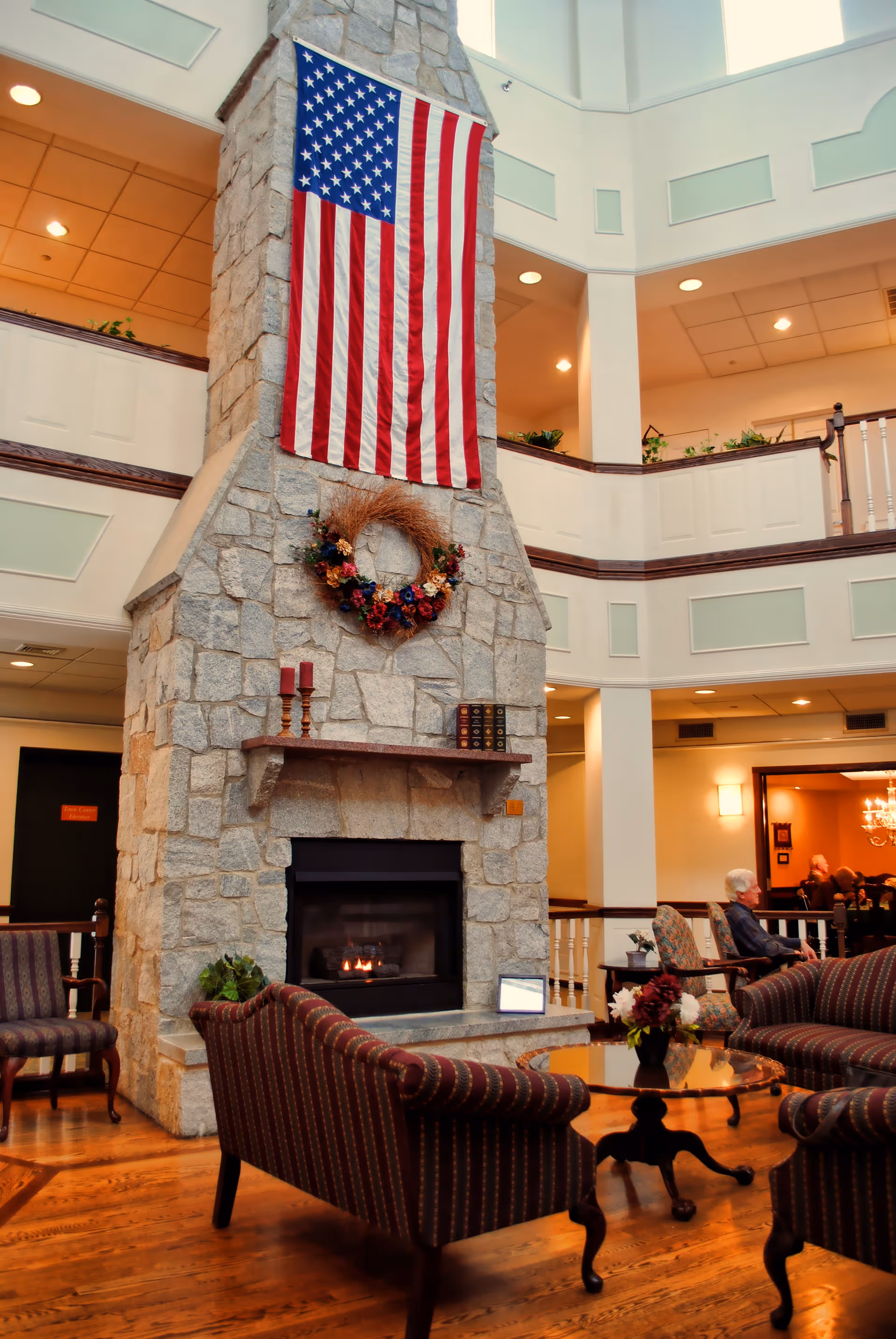 A spacious senior living facility common area with a tall stone fireplace adorned with an American flag and a decorative wreath. The room features wooden floors, comfortable upholstered chairs and sofas arranged around a glass coffee table with a floral centerpiece. The area is well-lit with recessed lighting and has a second-floor balcony overlooking the space. Two elderly people are seated in the background near a dining area.