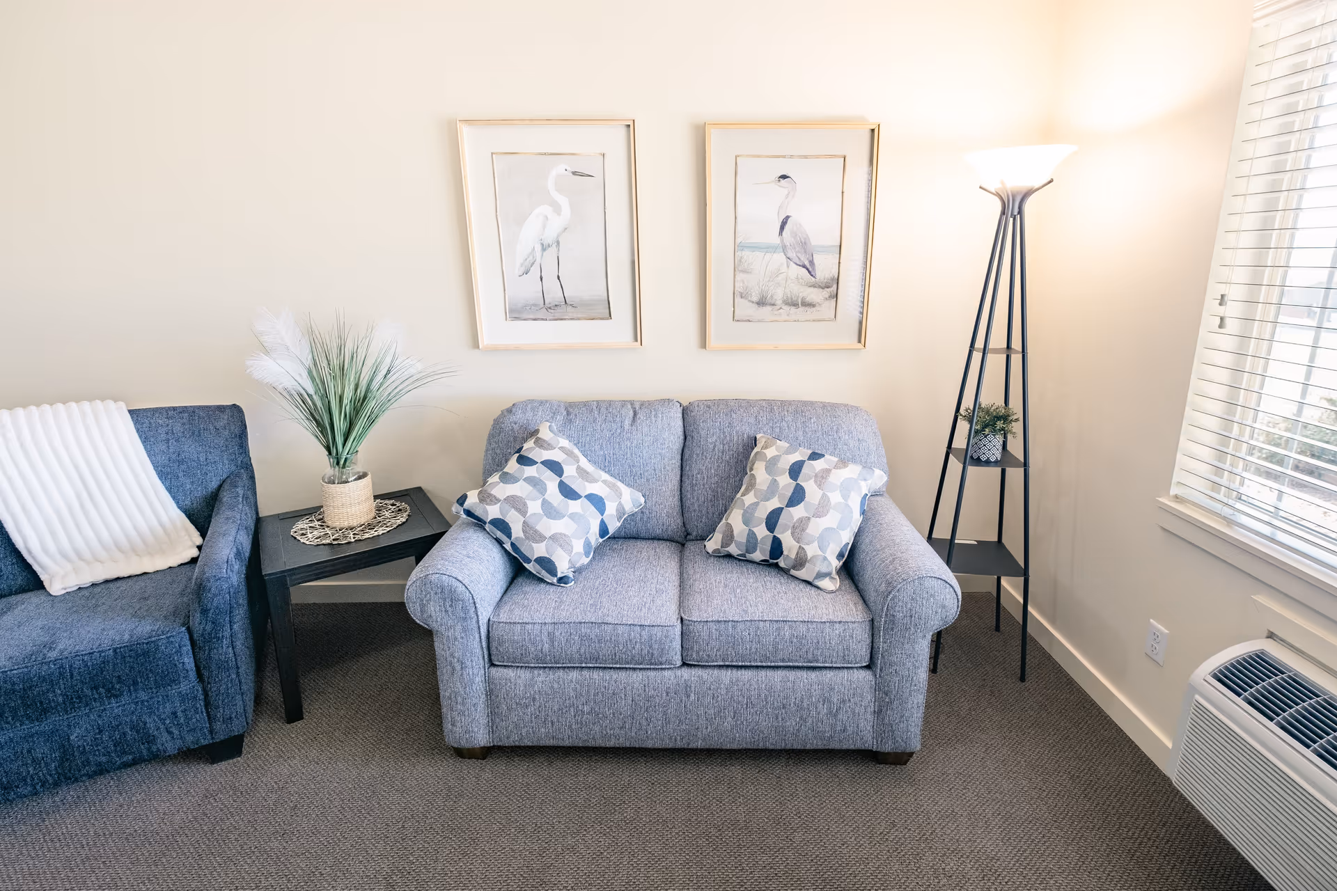 Small living room with a blue loveseat and armchair, patterned pillows, side tables, a tall floor lamp, framed bird artwork, and a window.