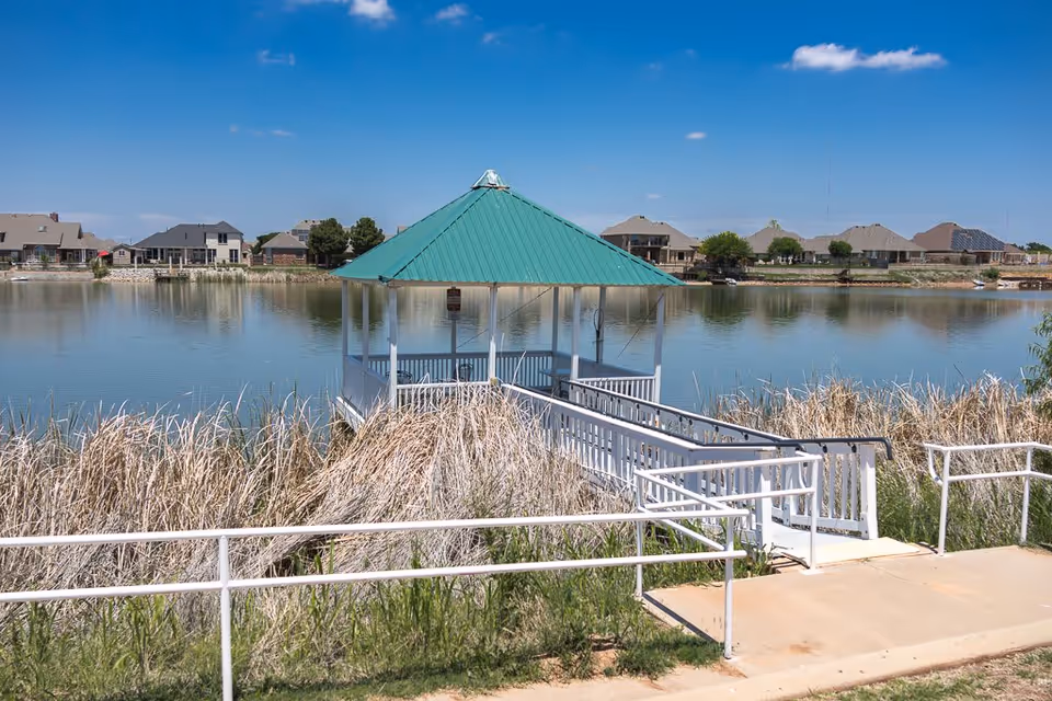 A white gazebo with a green roof at the end of a ramp overlooking a calm lake with houses on the far shore.
