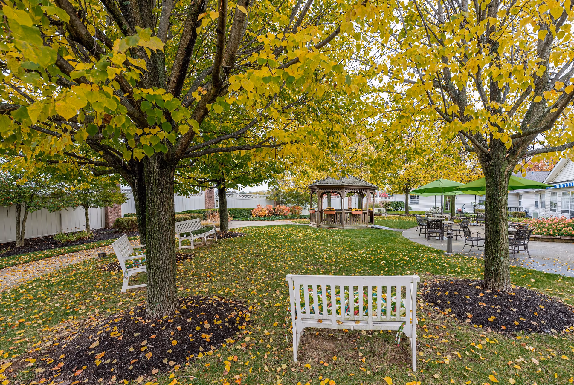 Outdoor garden area with several trees showing yellow and green leaves, white benches placed around the trees, a wooden gazebo in the background, and patio tables with green umbrellas near a white building.