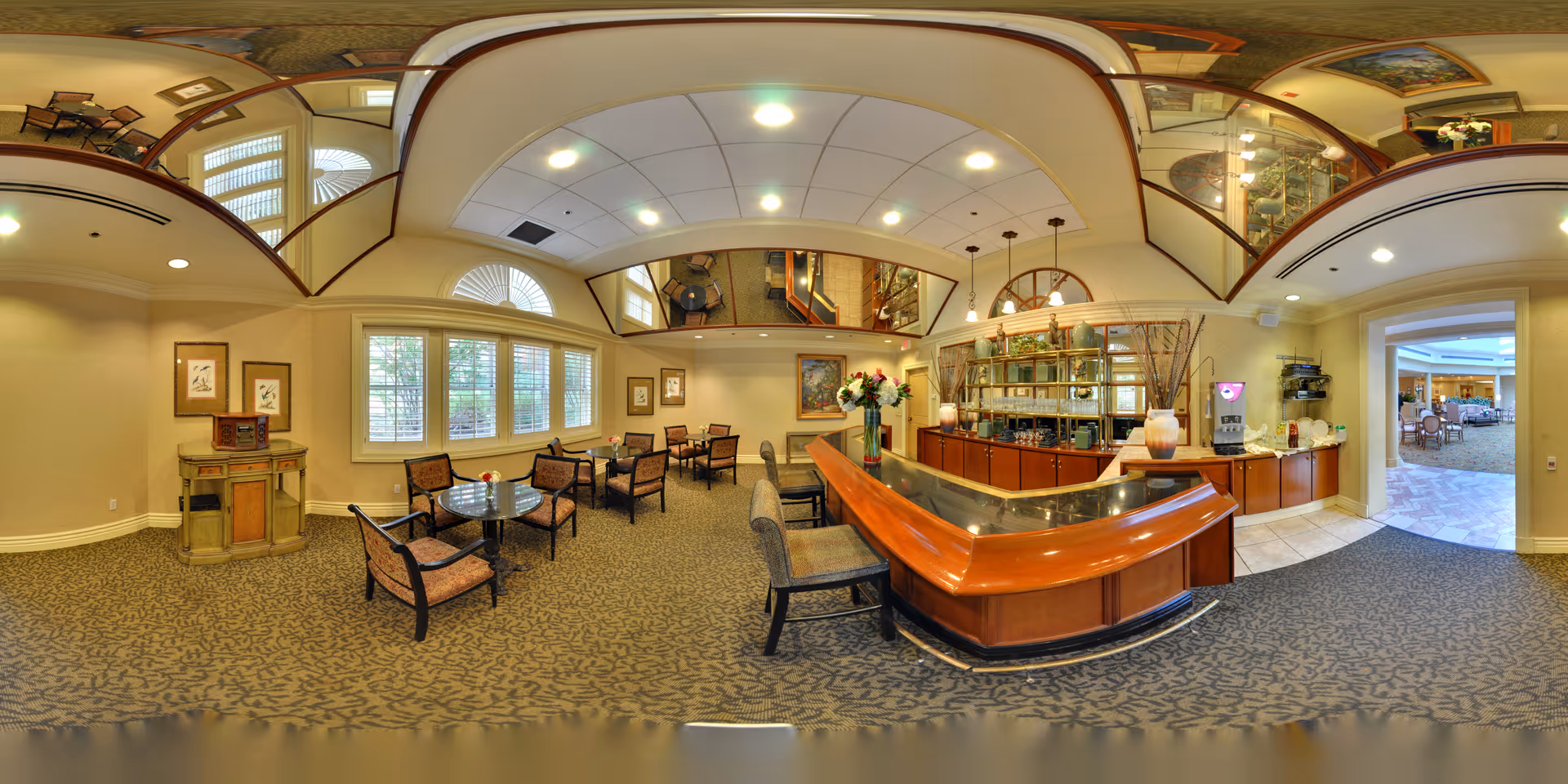 Panoramic interior view of a senior living facility lounge with a curved service counter, seating tables, and chairs.