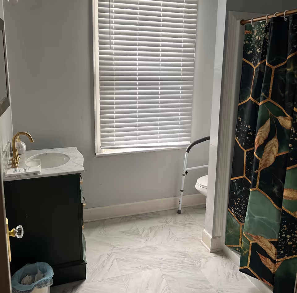 Bathroom with a window covered by white blinds, a dark green vanity with a white marble countertop and gold faucet, a trash can with a blue liner, a toilet with a safety frame, and a shower with a dark green and gold patterned curtain.