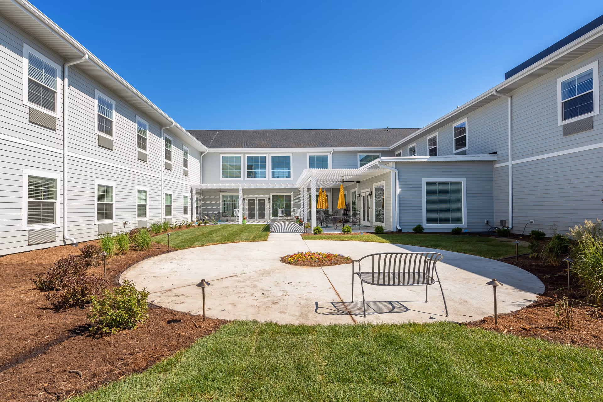 Exterior courtyard of a two-story senior living building with a circular paved area, a bench, landscaping, and seating under a pergola.