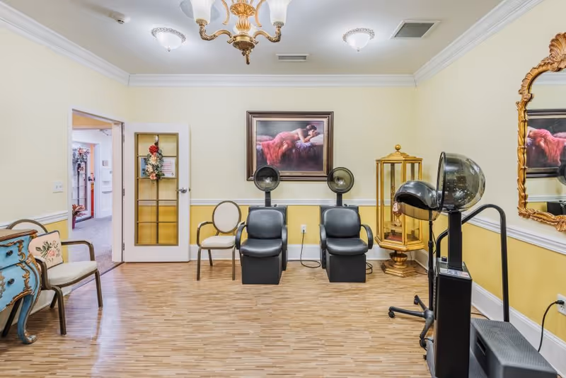 A room with two black salon hair dryers and chairs, a vintage blue dresser with a floral pillow, a beige chair, a gold-framed mirror, and a painting of a woman in a red dress lying down. The walls are painted yellow with white trim, and there is a chandelier and ceiling lights.