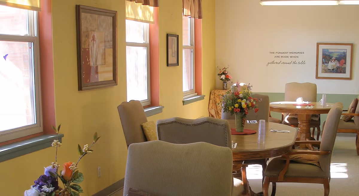 A bright dining room with round wooden tables and cushioned chairs. The walls are painted yellow with several windows letting in natural light. There are framed pictures and a wall decal that reads 'The fondest memories are made when gathered around the table.' Floral arrangements decorate the tables and a small side table in the corner.