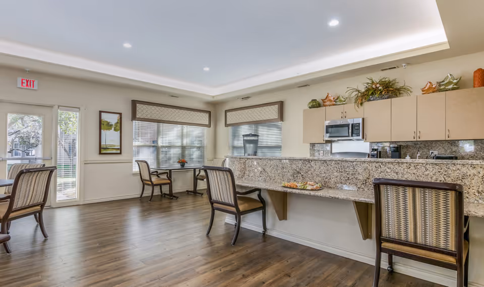 Bright communal dining area with a granite serving counter, chairs, tables, and large windows in a senior living facility.