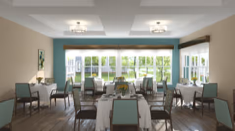 A bright dining room with multiple tables covered in white tablecloths, each surrounded by chairs with light blue upholstery. Large windows let in natural light and offer a view of greenery outside. The room has a modern ceiling with recessed lighting fixtures and wooden flooring.