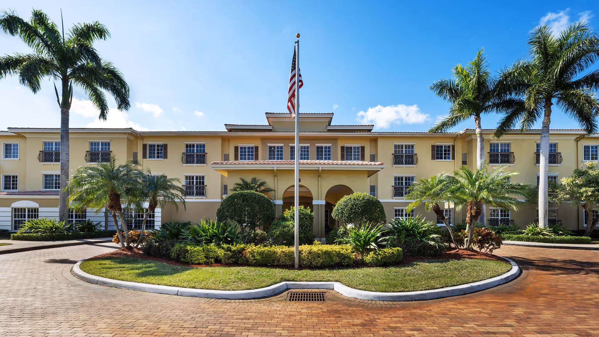 Front exterior view of a three-story senior living facility building with a circular driveway, landscaped greenery, palm trees, and an American flag on a flagpole in the center.