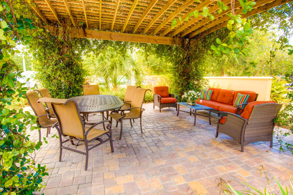 Outdoor patio area with a wooden pergola covered in climbing plants. The patio has a round metal table with six beige cushioned chairs on one side and a seating area with an orange cushioned sofa, two matching armchairs, a coffee table, and colorful striped pillows on the other side. The area is surrounded by greenery and trees.