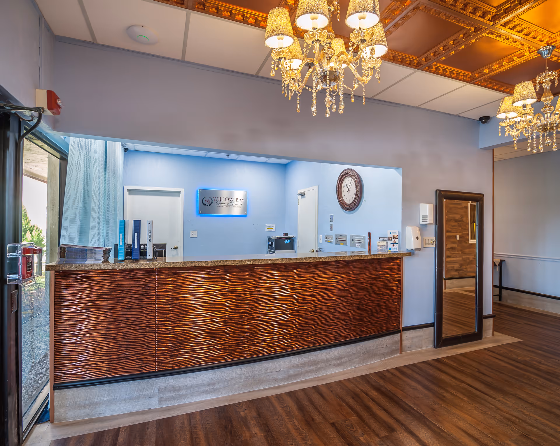 Reception area of Willow Bay Senior Resort featuring a wooden front desk with a granite countertop, a large wall clock, a mirror on the right wall, and elegant chandeliers hanging from a decorative ceiling. The background wall is light blue with a sign displaying the facility's name.