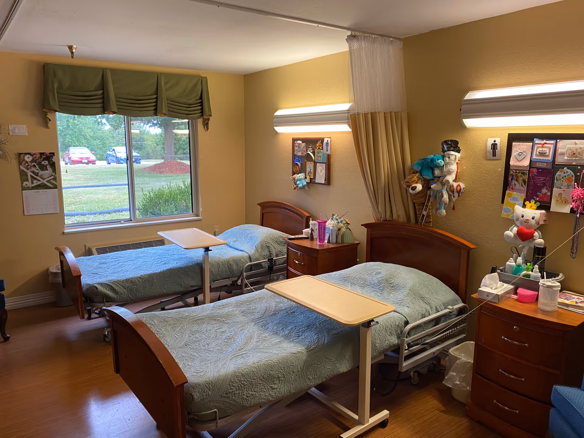 A room with two hospital-style beds covered with light blue quilts, each with an overbed table. The room has beige walls, wooden furniture including two nightstands, and a window with a green valance showing a view of parked cars and greenery outside. There are personal items and decorations on the walls and nightstands, including stuffed animals and a bulletin board.
