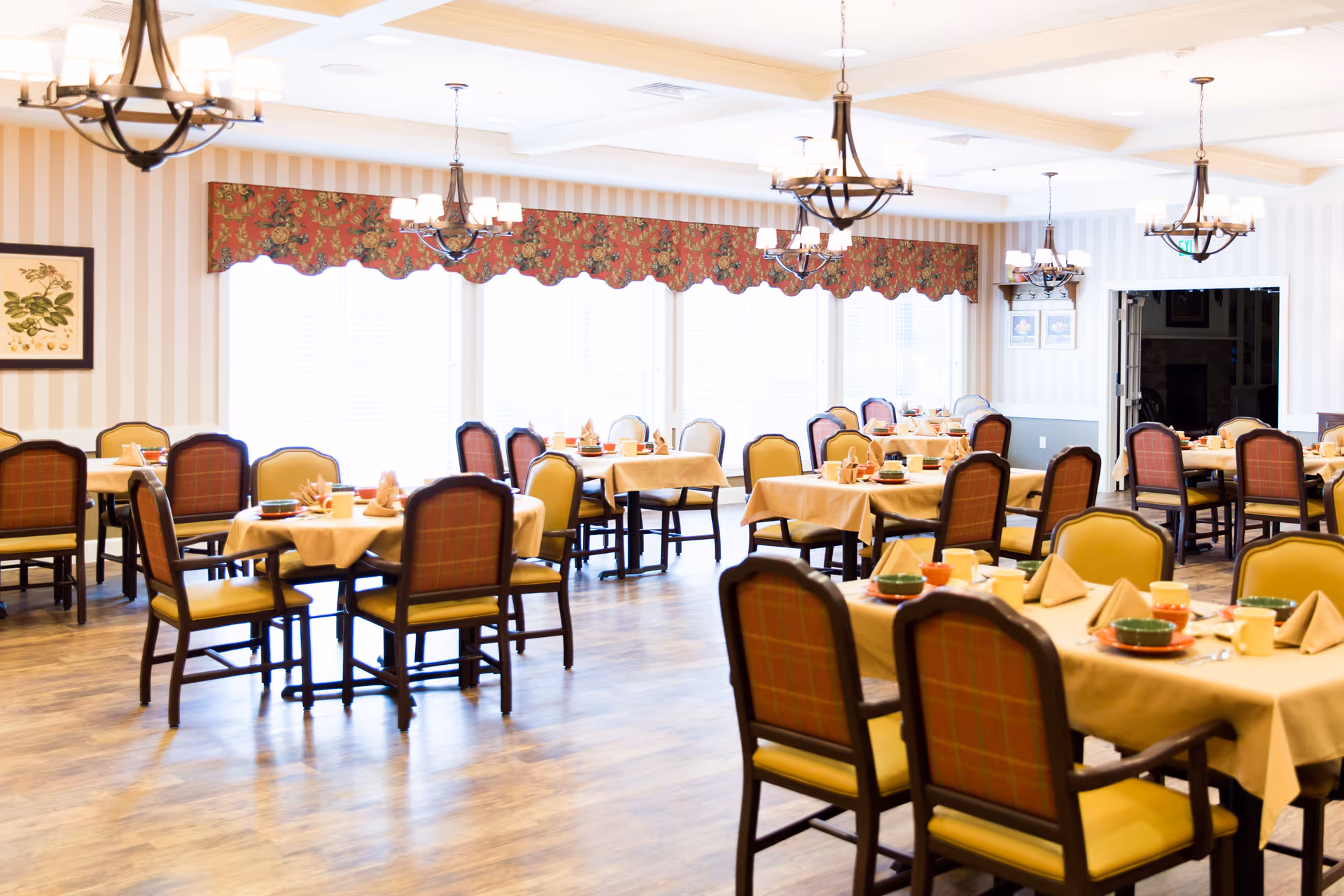 A bright and spacious dining room with multiple tables covered in beige tablecloths, each set with plates, bowls, cups, and folded napkins. The chairs have wooden frames with yellow and red plaid upholstery. Large windows with floral valances allow natural light to fill the room. Several chandeliers hang from the ceiling, and framed artwork decorates the striped walls.