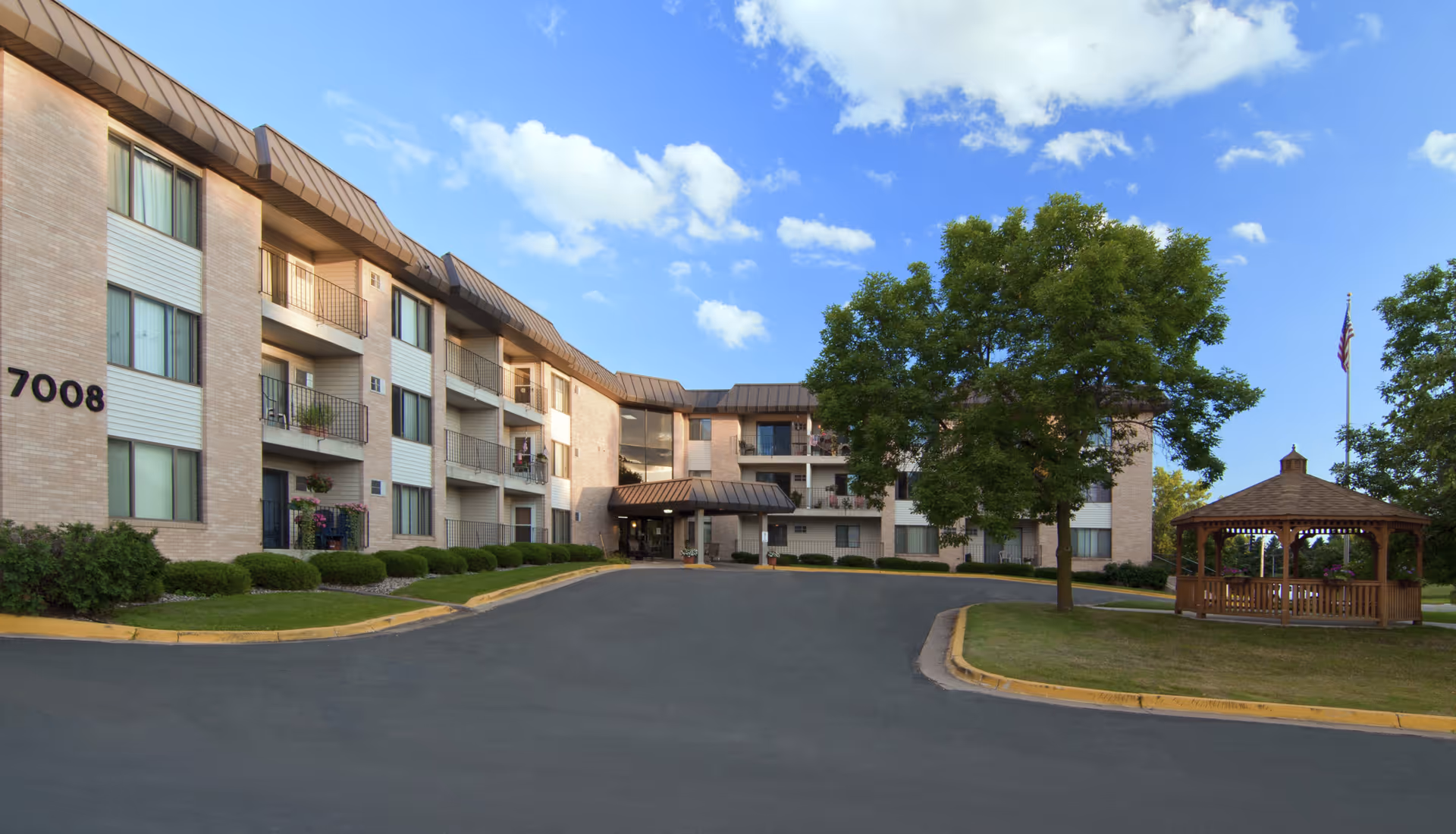 Exterior view of a three-story senior living facility building with balconies, a driveway, well-maintained landscaping including bushes and a large tree, and a wooden gazebo on the right side under a blue sky with scattered clouds.