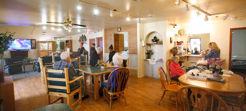 A common area in a senior living facility with several elderly residents sitting at tables and chairs. Some people are engaged in conversation while others are seated quietly. The room has wooden floors, a ceiling fan with lights, and a television mounted on the wall. There is a small sink area with shelves and flowers, and a woman is assisting another person near a vanity mirror. The atmosphere appears warm and communal.