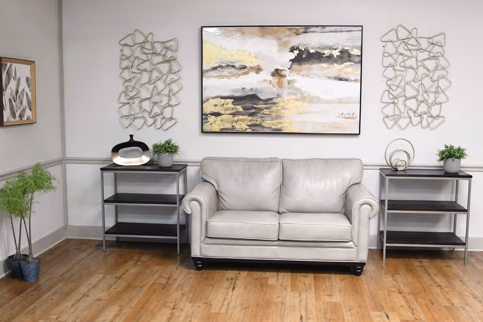 A cozy living room area with a light gray leather loveseat centered between two black metal and wood side tables. Each table holds decorative items including small green plants and modern sculptures. Above the loveseat hangs an abstract painting with gold, black, and white tones, flanked by two geometric metal wall art pieces. The room has light-colored walls with molding and a wooden floor.