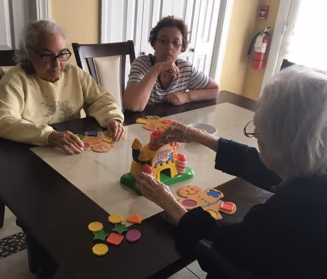 Three elderly women sitting around a table playing a colorful board game with various shaped pieces in a well-lit room with a window and a fire extinguisher on the wall.
