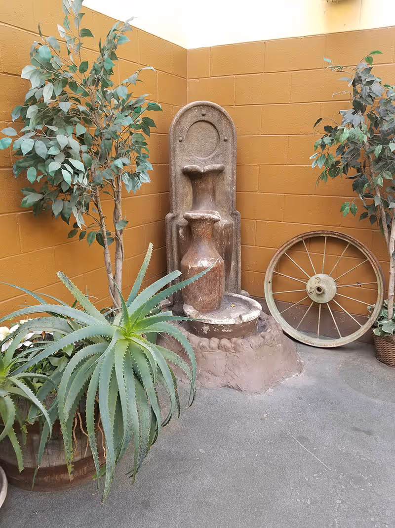 Decorative indoor corner with a tiered clay fountain, potted plants and a wooden wagon wheel against a tan cinderblock wall.