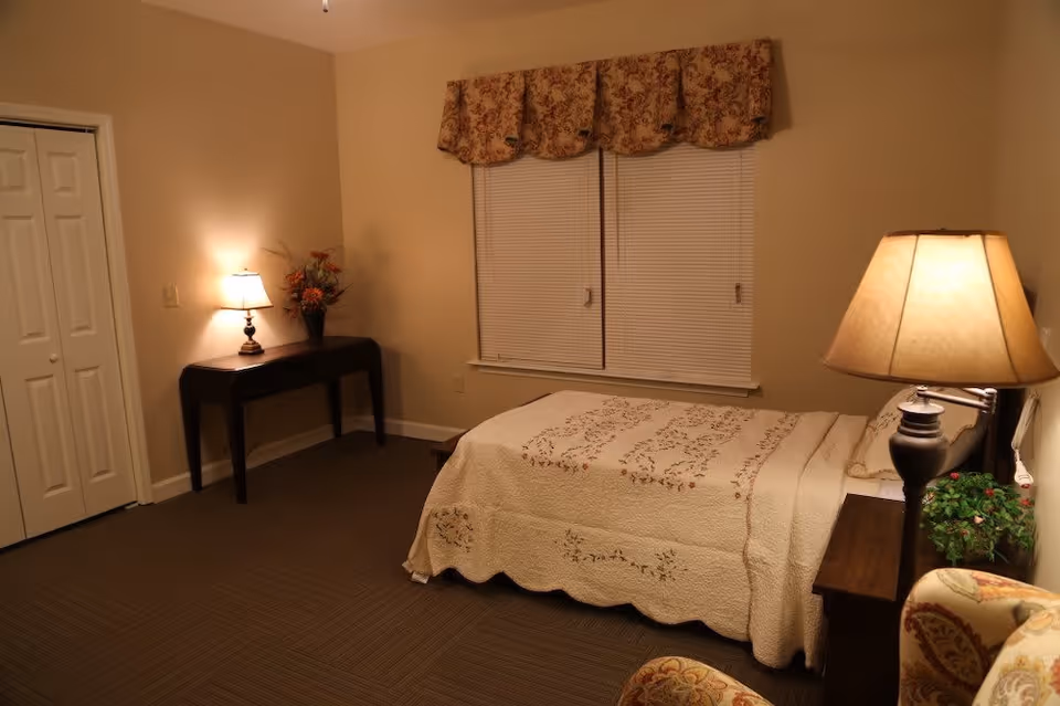 A cozy bedroom with a bed covered in a cream-colored quilt with floral embroidery. There is a wooden nightstand with a lamp and a small plant next to the bed. A window with closed blinds and a floral valance is on the wall behind the bed. A dark wooden table with a lamp and a flower arrangement is against the opposite wall. The room has beige walls and carpeted floor.