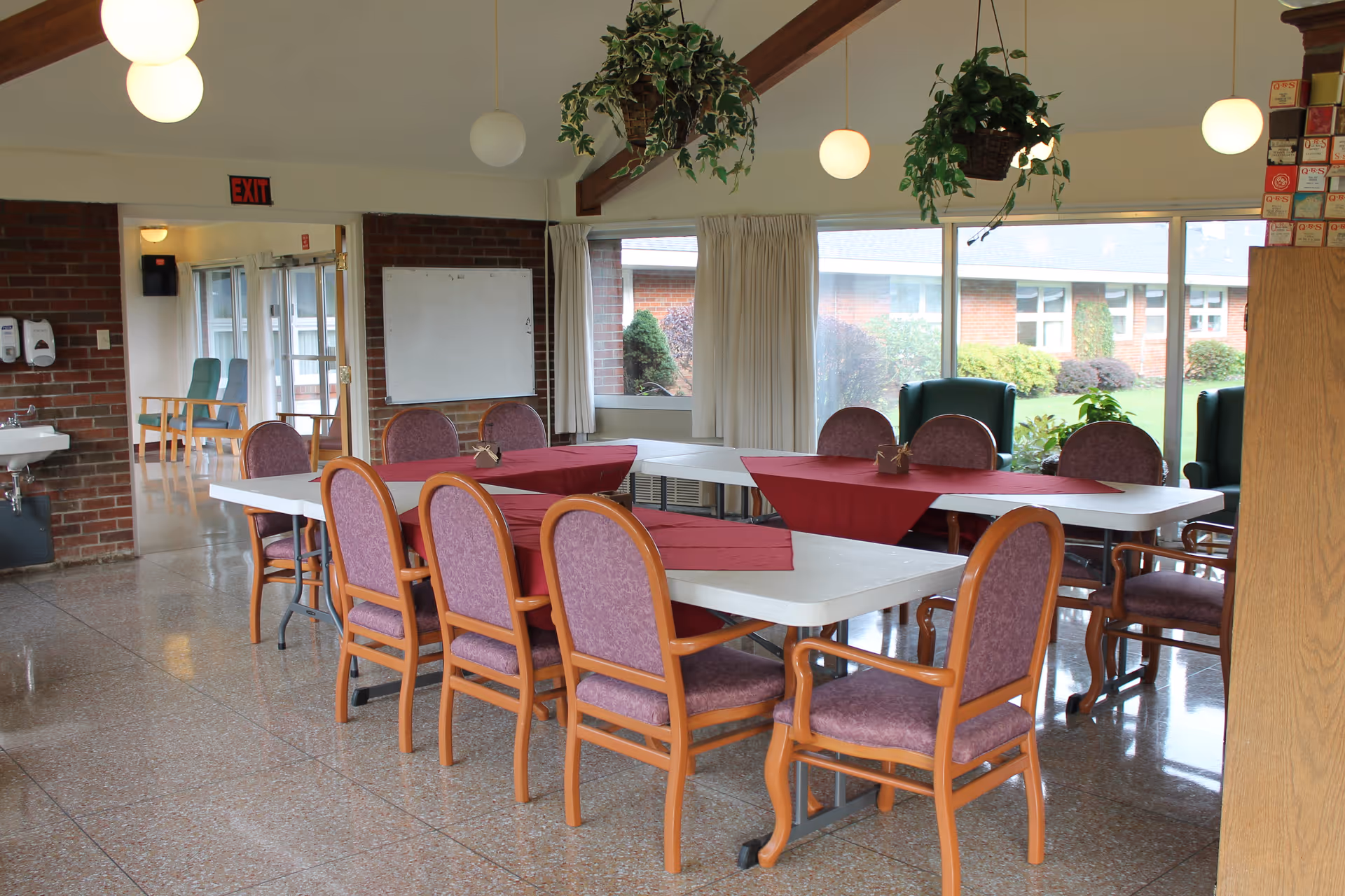 A dining area in a healthcare facility with two white rectangular tables arranged in an L-shape, covered with red table runners. There are multiple wooden chairs with purple cushions around the tables. The room has large windows with a view of a garden outside. Hanging plants and round pendant lights are suspended from the ceiling. A whiteboard is mounted on a brick wall near an exit door.