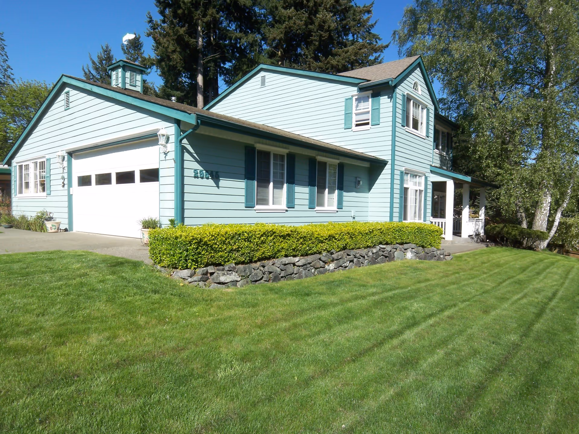 A two-story light blue house with teal trim and shutters, a white garage door, and a well-maintained green lawn with a stone border and bushes in front. Tall trees are visible in the background under a clear blue sky.