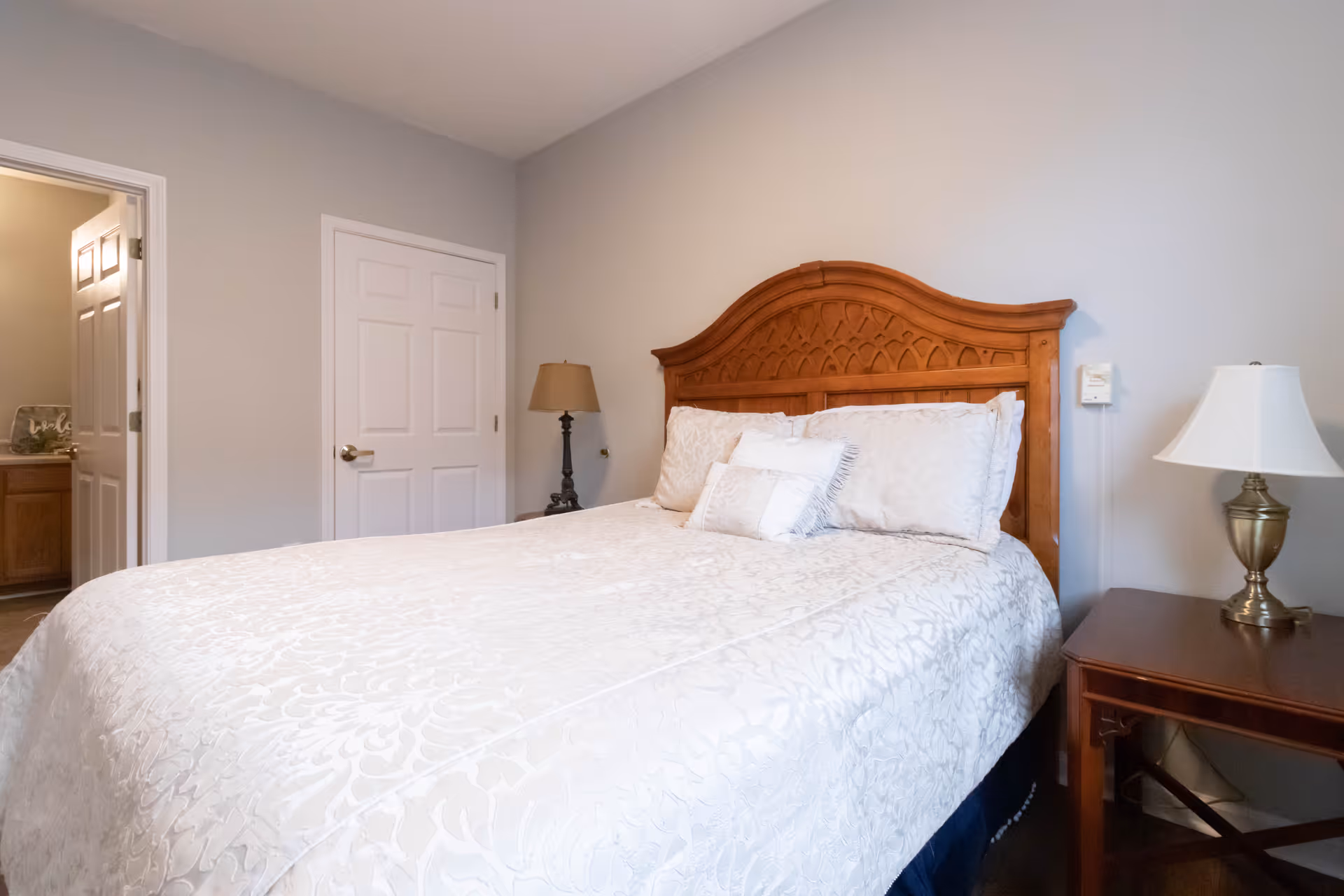 A bedroom with a wooden headboard bed covered in a white patterned bedspread and pillows. There is a wooden nightstand with a lamp on the right side of the bed and another lamp on a small table near a closed white door. An open door reveals a glimpse of a bathroom with a sink and countertop.