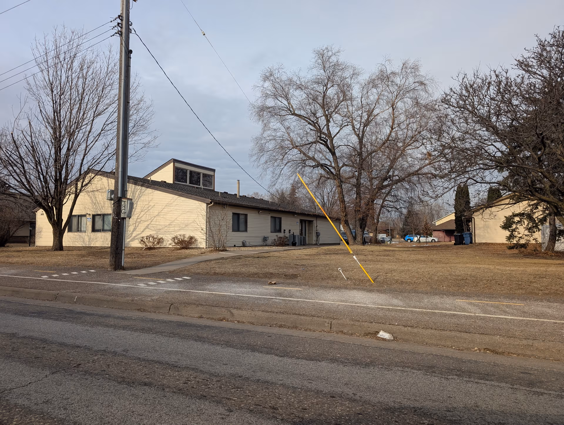 Exterior view of a single-story beige building with a sloped roof, surrounded by leafless trees and a dry grassy area. A utility pole with wires is visible in the foreground near a paved road.