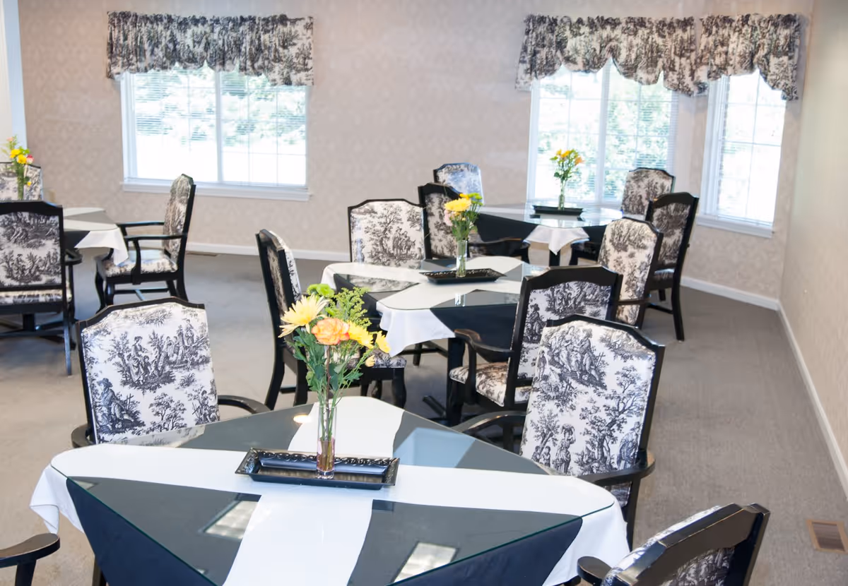 Bright assisted-living dining room with multiple tables, patterned chairs, and floral centerpieces near large windows.