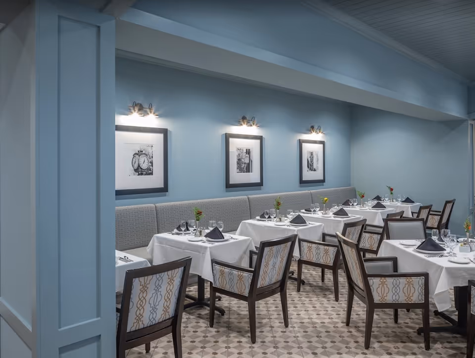 Dining area with multiple tables covered in white tablecloths, each set with black napkins, glassware, and small flower vases. The room has light blue walls with three black and white framed pictures illuminated by wall-mounted lights. Chairs with patterned upholstery surround the tables, and the floor has a patterned tile design.