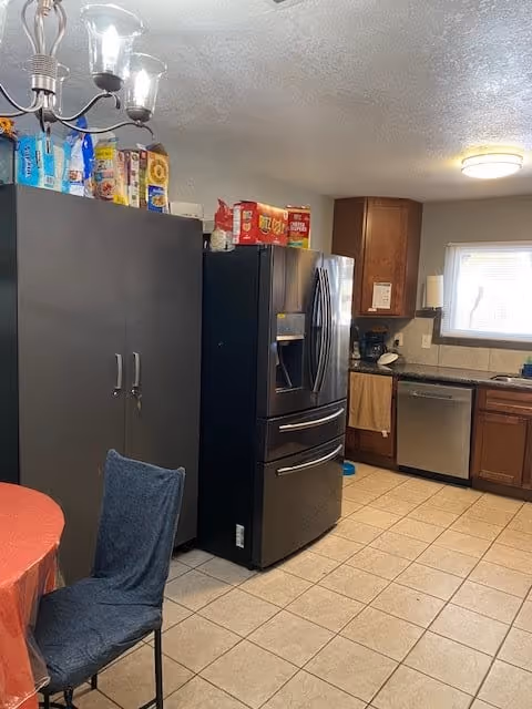A kitchen with tiled floor, a black refrigerator, wooden cabinets, a dishwasher, and a window above the sink. There is a gray storage cabinet with various food items on top, a round table with a red tablecloth, and a chair with a blue cover. A ceiling light fixture with three glass shades is visible.