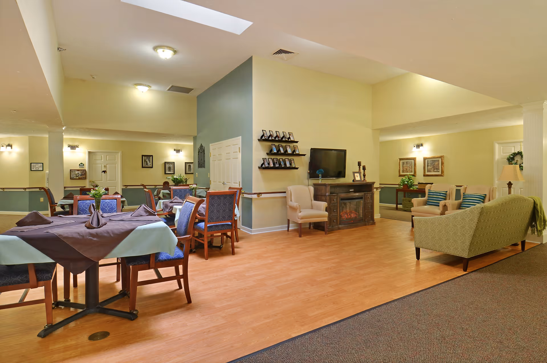 Interior view of a senior living facility common area with a dining section on the left featuring tables covered with tablecloths and chairs, and a living area on the right with armchairs, a loveseat, a TV mounted above a fireplace, and framed pictures on the walls. The space has wooden flooring, soft lighting, and a calm, welcoming atmosphere.