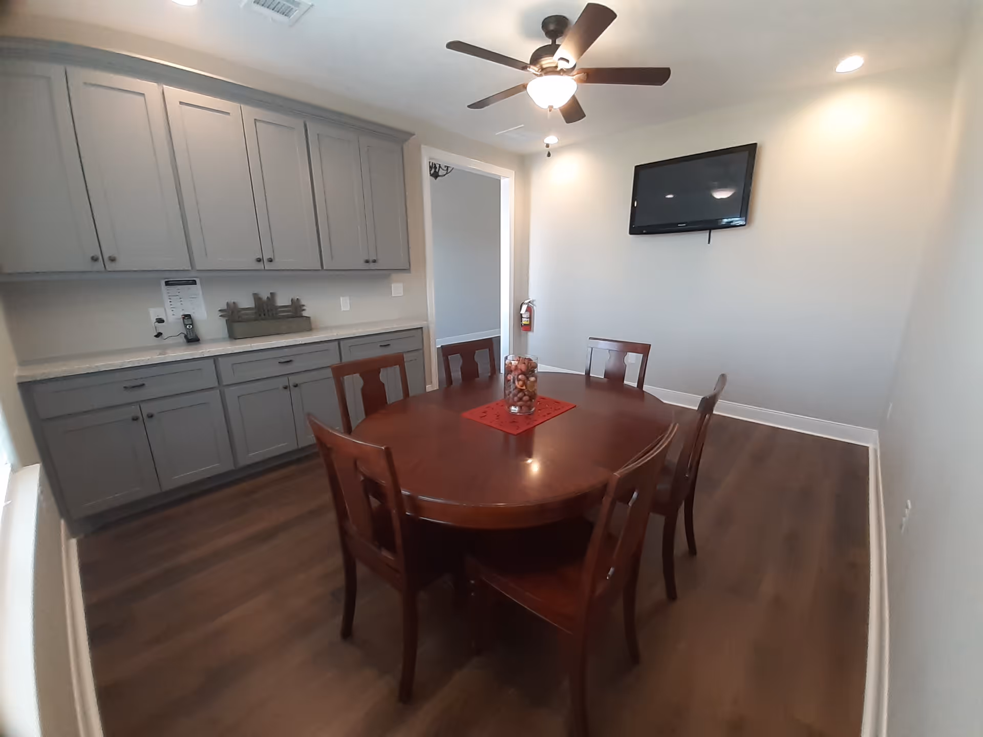 A dining room with a round wooden table surrounded by six matching wooden chairs. On the table is a glass vase filled with decorative items placed on a red placemat. The room has gray cabinets along one wall, a ceiling fan with a light, recessed lighting, and a wall-mounted flat screen TV. The floor is wood, and there is a fire extinguisher mounted on the wall near an open doorway.
