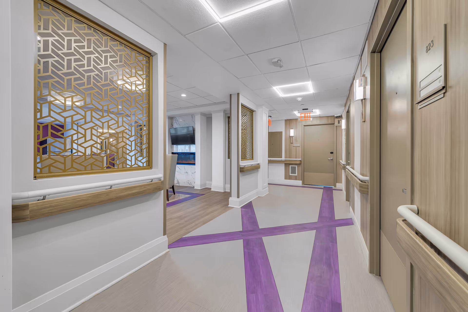 A clean, modern hallway in The Veranda Assisted Living facility featuring beige doors with room numbers, decorative gold geometric window panels, wooden handrails, and a floor with intersecting purple lines. The hallway is well-lit with ceiling lights and has an exit door at the end.