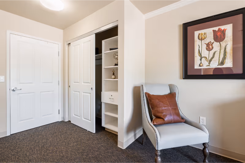 A corner of a room featuring a white door and a closet with sliding doors partially open, revealing shelves inside. Next to the closet is a light gray upholstered chair with a brown leather pillow. Above the chair hangs a framed artwork of three tulips on a beige wall. The floor is carpeted in a dark pattern.