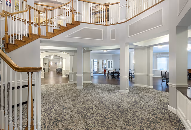 Spacious interior of a senior living facility featuring a carpeted area with a wooden staircase and white railings leading to an upper floor. The room has multiple columns and open spaces with tables and chairs in the background, illuminated by ceiling lights and natural light from windows.