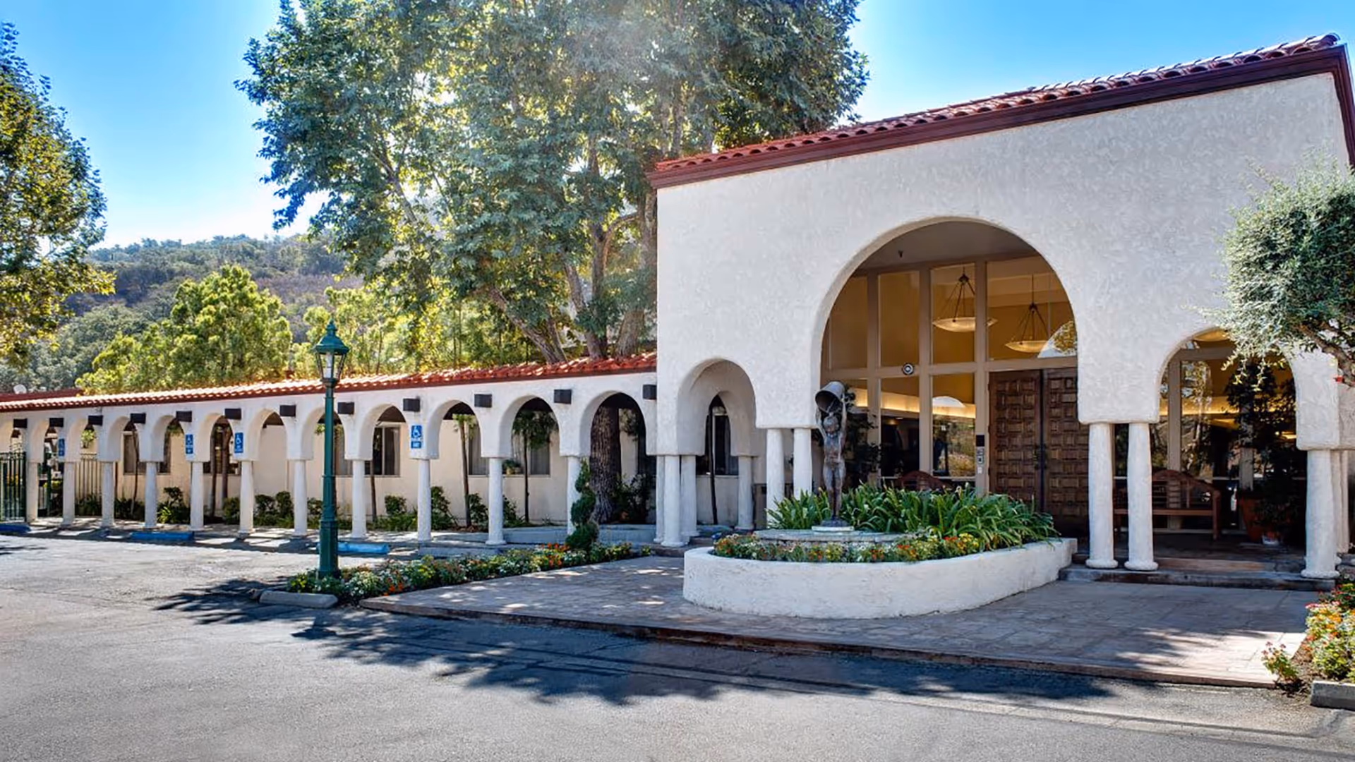 Exterior view of Silverado Calabasas Memory Care Community featuring a white stucco building with red tile roof, arched entryways, and a landscaped area with plants and a sculpture in front. Trees and hills are visible in the background under a clear blue sky.