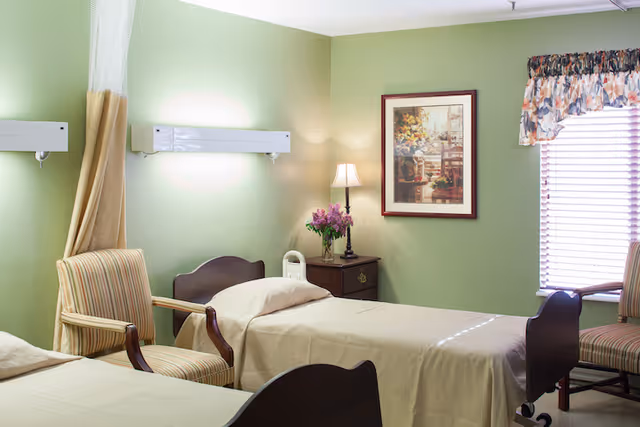 A room with two single beds covered in beige bedspreads, each accompanied by a striped armchair. The walls are painted light green, and there is a nightstand between the beds with a lamp and a vase of purple flowers. A framed floral painting hangs on the wall, and a window with floral valance and blinds lets in natural light.