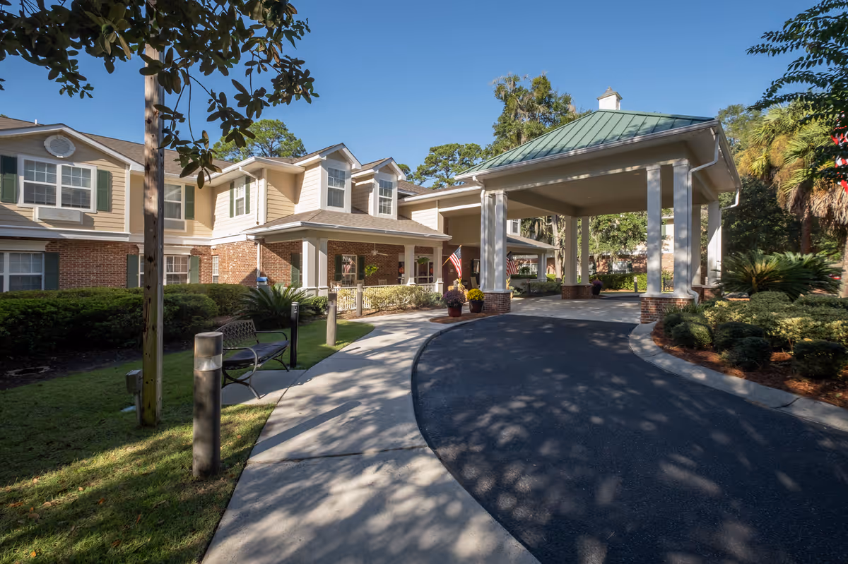 Front entrance of a two-story senior living building with a covered porte-cochère, curved driveway, benches, and landscaped grounds.