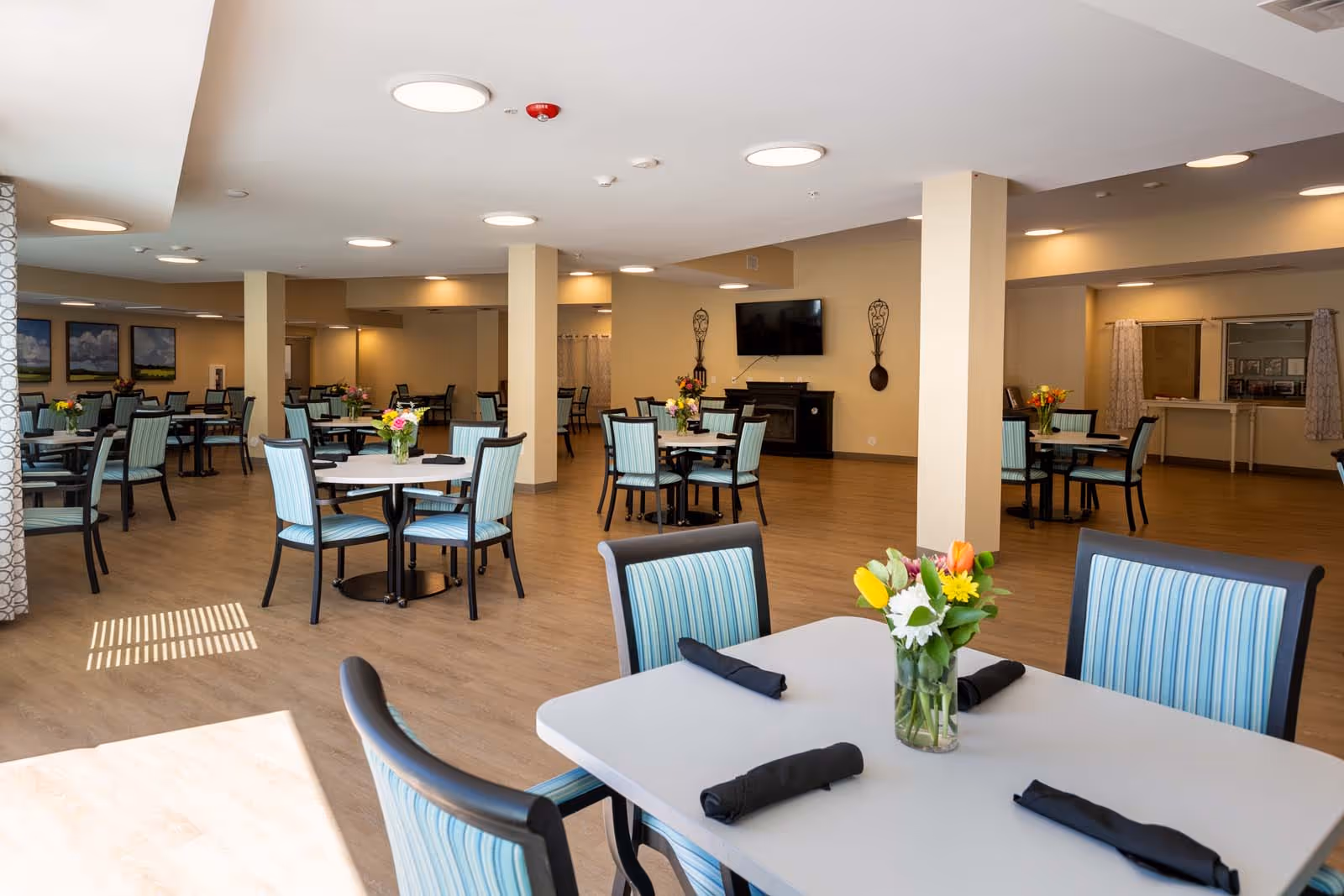 A spacious dining room with multiple round tables and chairs featuring blue striped upholstery. Each table has a vase with colorful flowers and black napkins neatly placed. The room has wooden flooring, beige walls, ceiling lights, a wall-mounted TV, and decorative wall sconces. Large windows allow natural light to brighten the space.