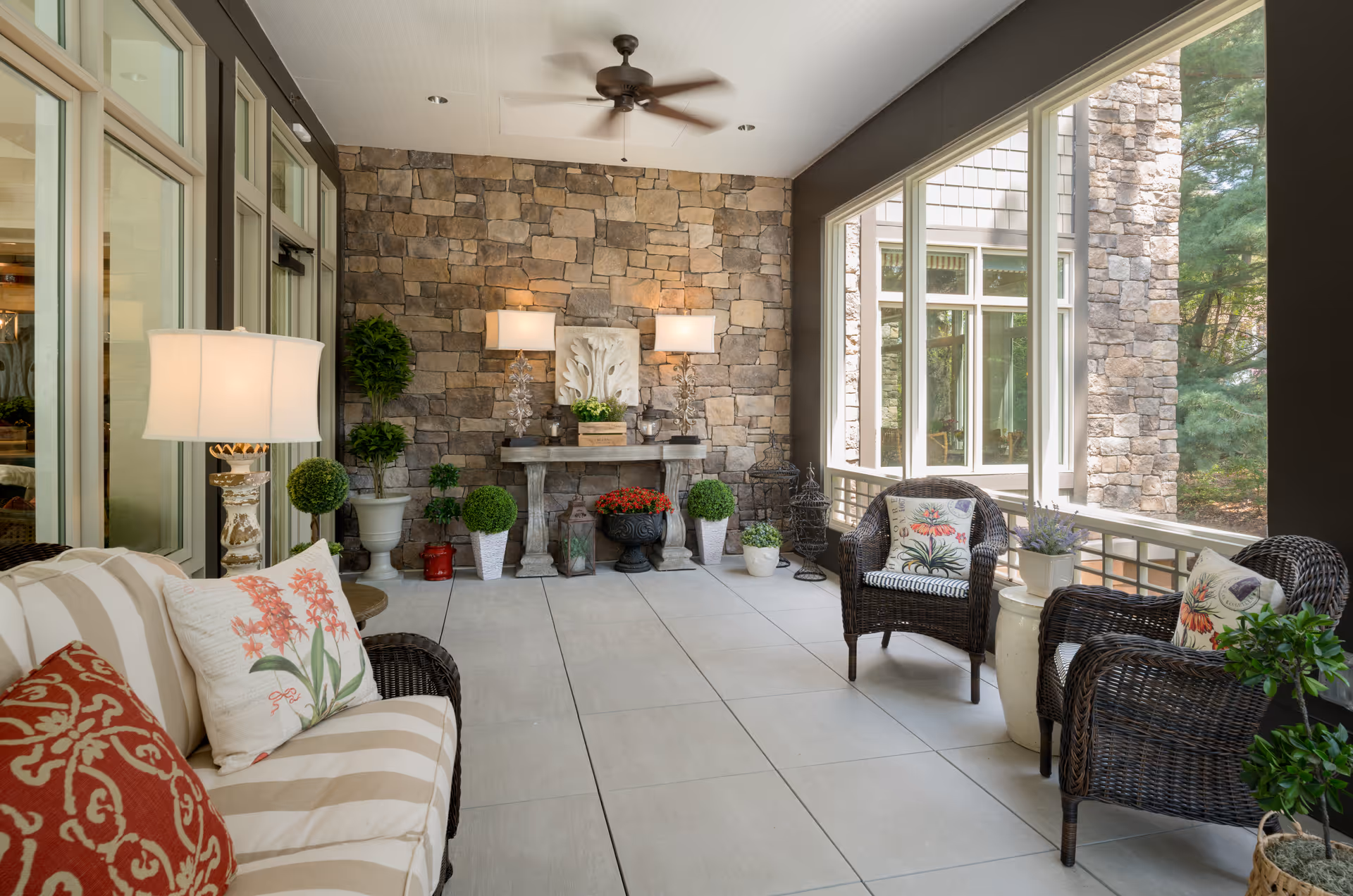 A cozy enclosed patio area with a stone wall, ceiling fan, and large windows. The space is furnished with a striped cushioned sofa with floral pillows, two wicker chairs with floral cushions, a side table with a lamp, and various potted plants and decorative items arranged on the floor and a stone console table.