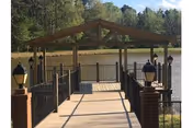 Wooden dock with a covered pavilion and lamp posts extending over a lake with trees in the background.
