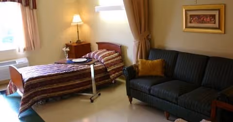 A cozy room featuring a single bed with striped bedding, a bedside table with a lamp, a small overbed table with a plate and cup, and a dark green upholstered couch with a mustard-colored pillow. The room has beige walls, a window with curtains, and a framed picture on the wall.