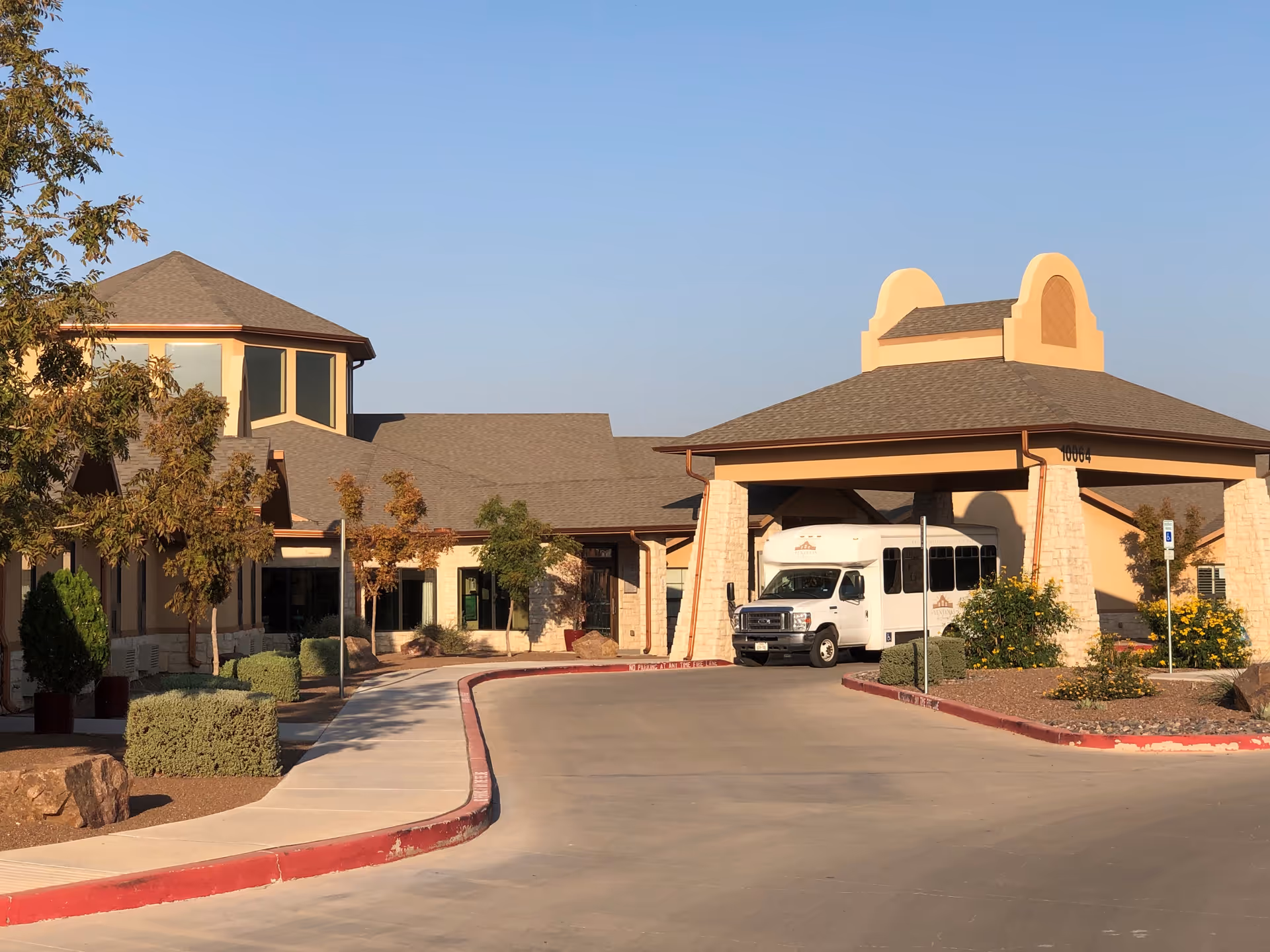 Exterior view of a senior living facility named Las Ventanas de Socorro with a covered entrance, a white shuttle bus parked under the canopy, landscaped bushes, trees, and a clear blue sky.