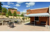 Outdoor patio area at Townview Health And Rehabilitation Center featuring metal tables and chairs on a paved surface, a brick building with a covered entrance, and a landscaped stone retaining wall with small trees and shrubs under a blue sky with some clouds.