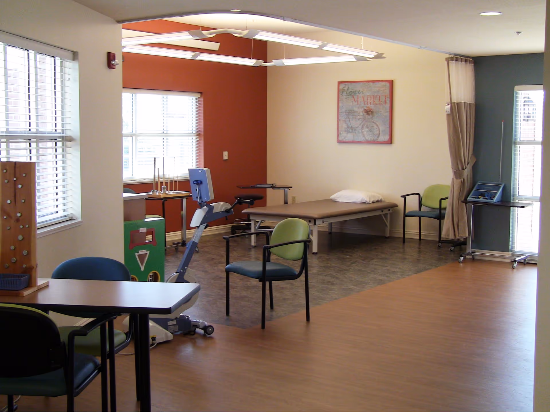 A spacious room with wooden flooring and large windows letting in natural light. The room contains several chairs, a table, a stationary exercise bike, a therapy or massage table with a pillow, and some therapy or activity equipment. The walls are painted in warm tones with a framed picture hanging on one wall.