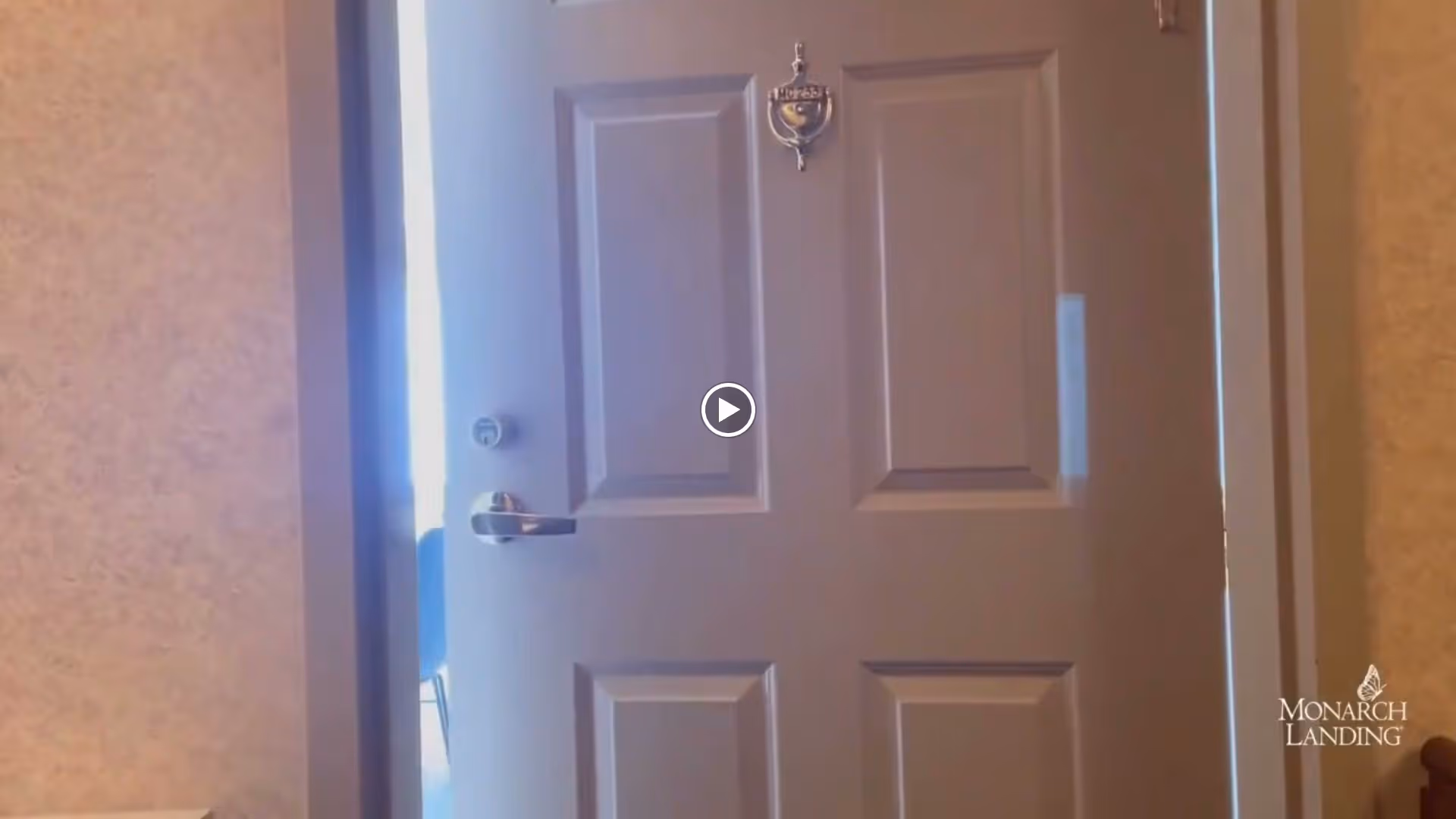 Partially open door with a silver handle and a door knocker, revealing a glimpse of a chair and a bright room beyond. The door has a beige frame and is set against a textured beige wall.