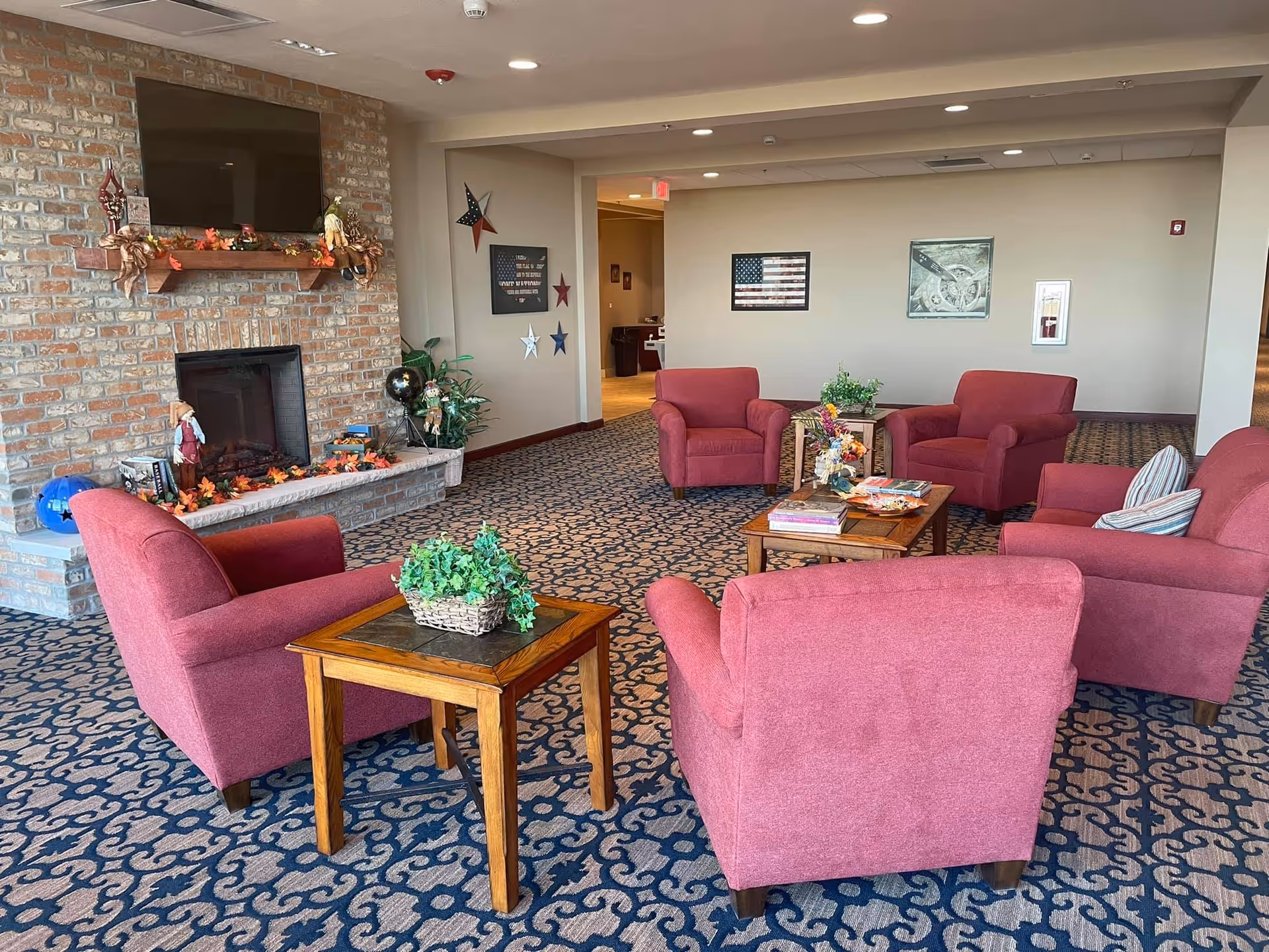A cozy living room area with five red upholstered armchairs arranged around two wooden coffee tables. The tables have decorative plants and magazines on them. There is a brick fireplace with a wooden mantel decorated with autumn-themed items and a flat-screen TV mounted above it. The room has patterned carpet and beige walls adorned with patriotic decorations and framed pictures.