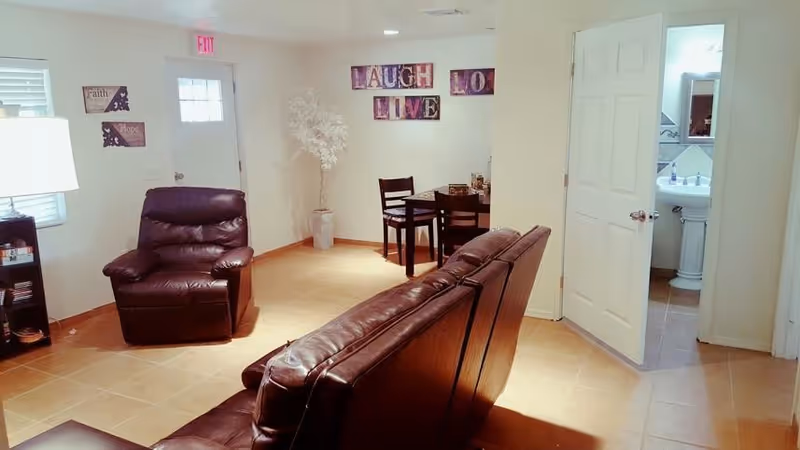 A cozy living room area with brown leather seating including a sofa and an armchair. There is a small dining table with four chairs in the corner near a white door. Wall decorations include framed words 'LAUGH' and 'LIVE' and smaller plaques with the words 'Faith' and 'Hope'. A white artificial tree stands in the corner. The floor is tiled and there is a doorway leading to a bathroom with a pedestal sink and mirror visible.