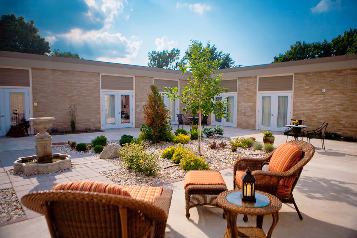 Sunny enclosed courtyard with wicker chairs, a small fountain, and landscaped gravel beds in front of ground-level apartment doors.