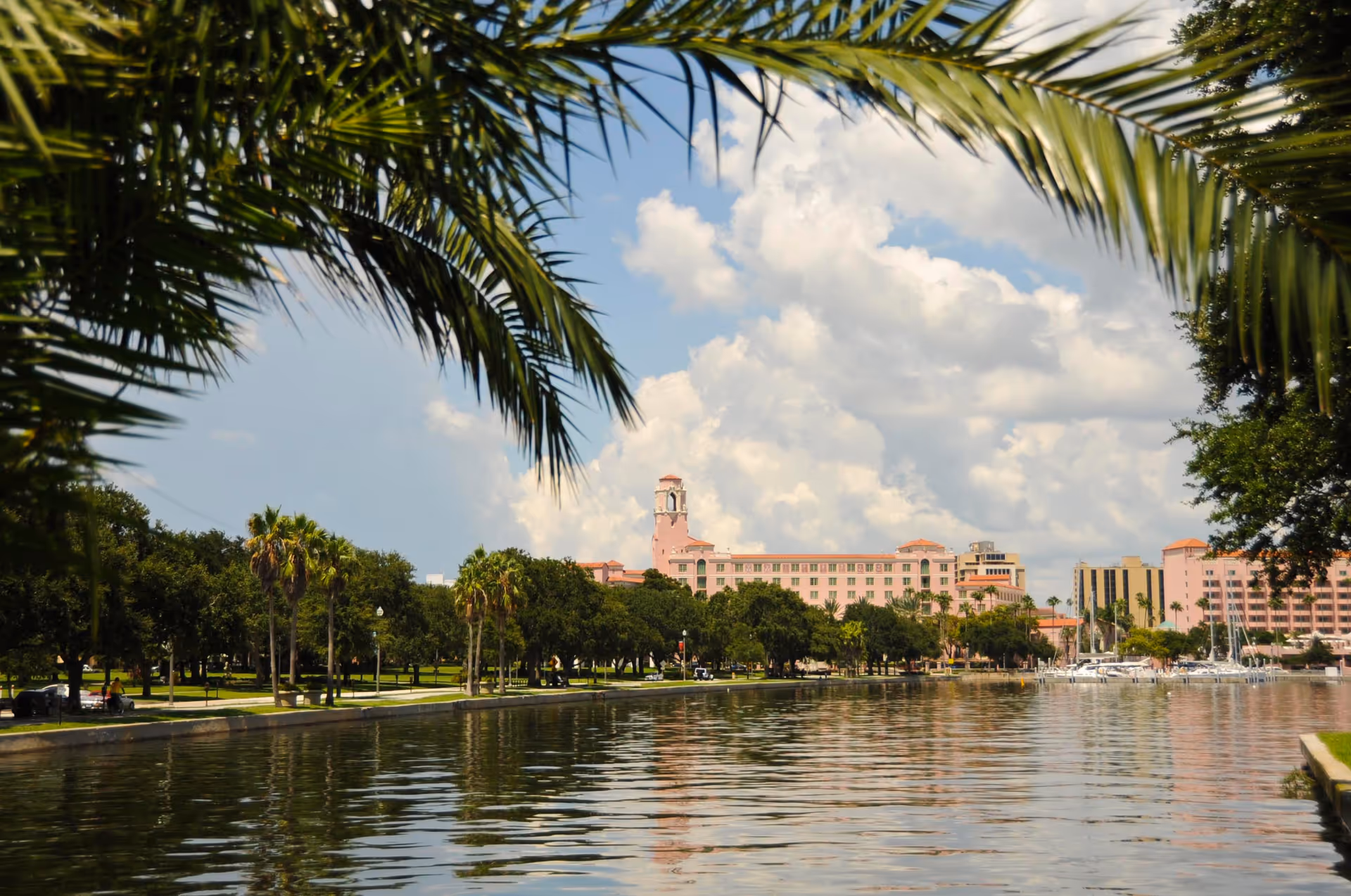 View of a waterfront park with palm trees and other greenery along the shore, calm water reflecting the sky, and a large pink building with a tower in the background under a partly cloudy sky.